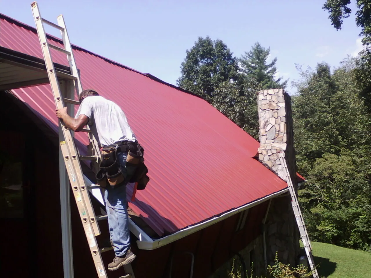 Roofer on a ladder beside a red metal roof and stone chimney — Soffit & Fascia Repair by Roofing Brothers in Deridder